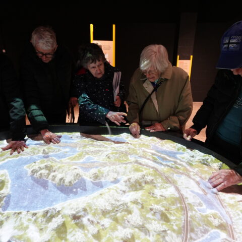 Photo shows the group of 5 people are stood around a sand pit, which reflects an interactive scene on the sand. The group are feeling the sand and interacting with the map.