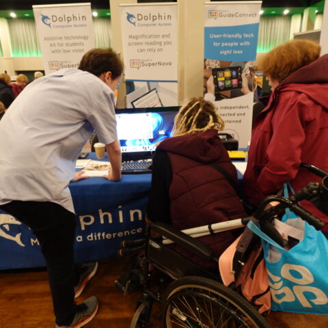 A stall holder at Sight Village North East 2024 in Leeds, showcasing tech to a member, who is in a wheelchair, and her friend. There are Dolphin Computer Access banners in the background.