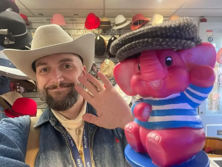 A man in a cream cowboy hat and MySight York lanyard takes a smiling selfie in a hat shop, waving beside a large pink toy elephant wearing a tweed flat cap, with rows of hats on display behind them.
