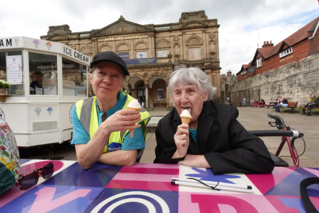 A MySight York volunteer in a hi-vis vest and an older woman enjoy ice cream cones at a colourful table outside York Art Gallery, with an ice cream van behind them, a folded white cane on the table and a rollator parked alongside.