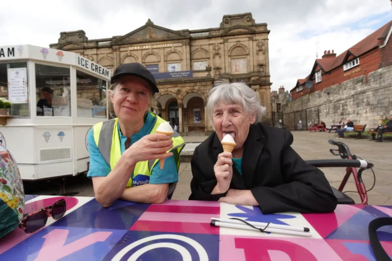 A MySight York volunteer in a hi-vis vest and an older woman enjoy ice cream cones at a colourful table outside York Art Gallery, with an ice cream van behind them, a folded white cane on the table and a rollator parked alongside.