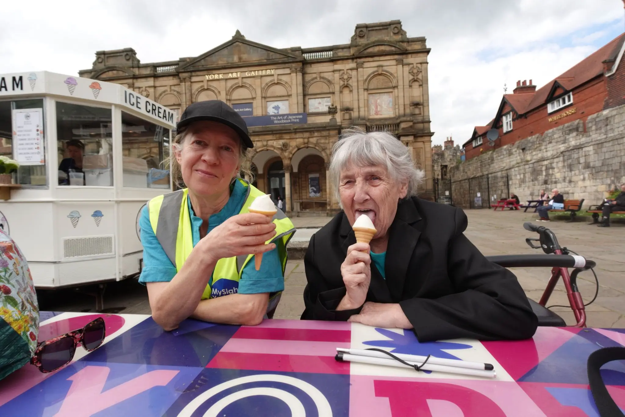 A MySight York volunteer in a hi-vis vest and an older woman enjoy ice cream cones at a colourful table outside York Art Gallery, with an ice cream van behind them, a folded white cane on the table and a rollator parked alongside.