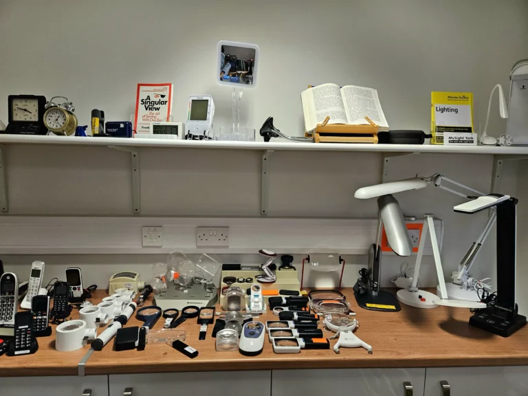 A worktop and shelf displaying assistive equipment: large-button phones, handheld and stand magnifiers, talking clocks, a video magnifier, task lamps, and a book on a wooden reading stand.