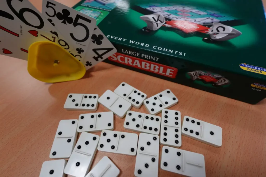 Dominoes spread across a table beside playing cards in a card holder and a large print Scrabble box.