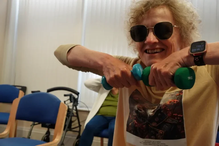 A smiling woman in dark glasses holds a pair of small dumbbells out in front of her at a seated exercise class.