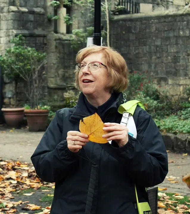 A woman in a dark jacket holds a large golden leaf and smiles as she looks up, standing on a leaf-strewn path beside old stone walls.
