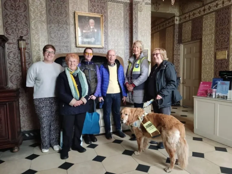 Six people and a golden retriever guide dog in a hi-vis harness pose in the ornately wallpapered hallway of a historic house, beside a large fireplace and portrait.