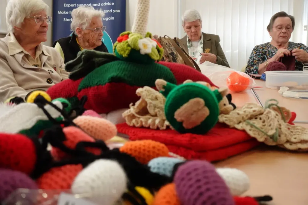 Four women knit and crochet around a table, with colourful finished pieces piled in front of them.