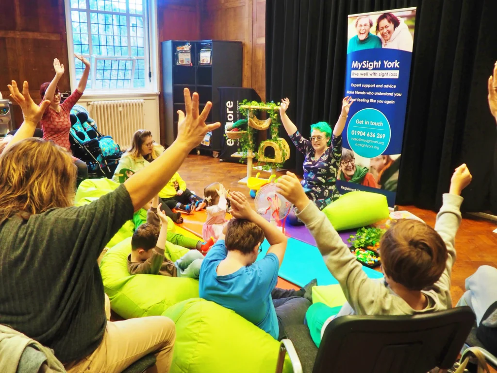 Children and adults raise their hands in the air during a song at a MySight York family session, sitting on green beanbags beside a MySight York pull-up banner.