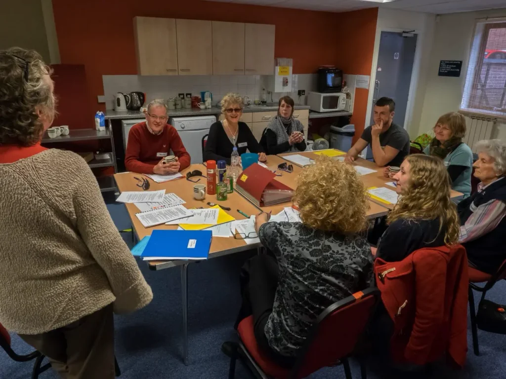 A group of around ten people sit around tables in a community room kitchen, with paperwork, folders and tea in front of them, listening to a woman standing to speak.