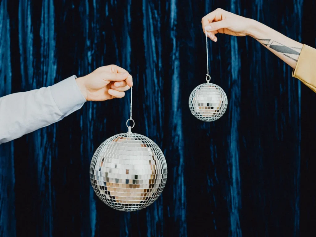 Two outstretched arms hold a large and a small mirrored disco ball on strings against a dark blue velvet curtain.