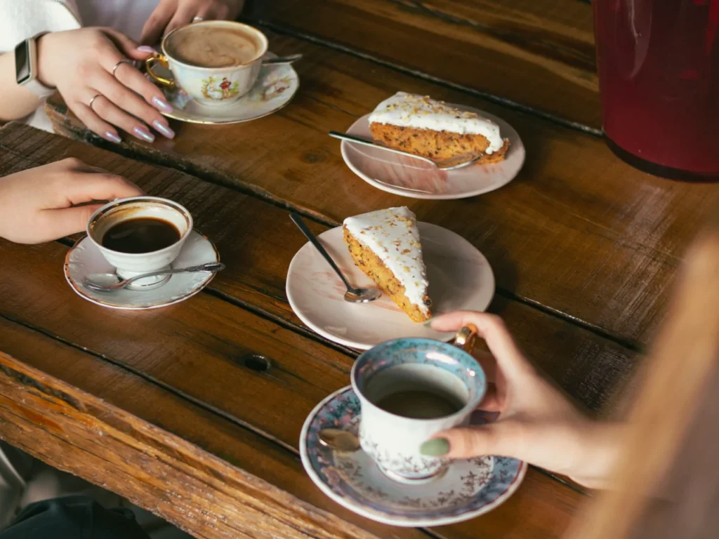 Overhead view of friends' hands around a wooden café table with vintage china cups of coffee and slices of iced carrot cake.