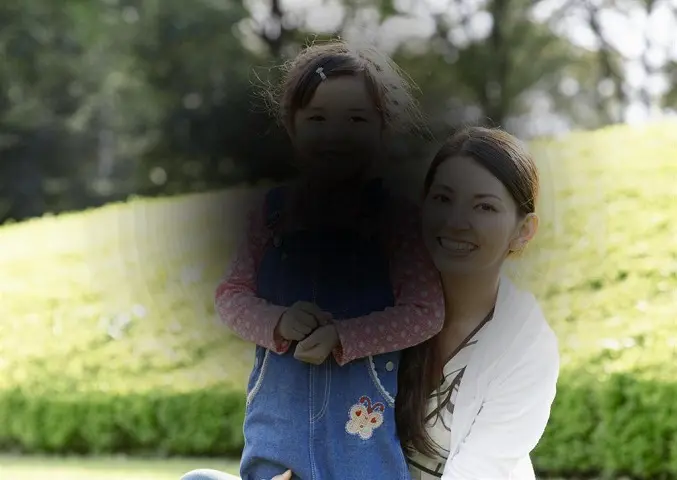 A woman and young girl smile in a sunny park, but the girl's face and the centre of the image are darkened and blurred — simulating the loss of central vision caused by conditions like macular degeneration.