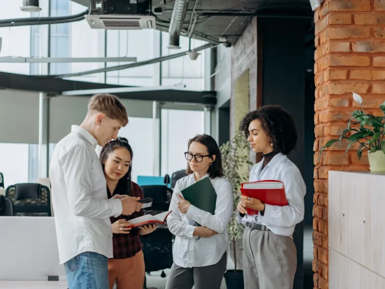 Four young colleagues stand chatting in a modern open-plan office, one holding an open notebook while two others hold folders.