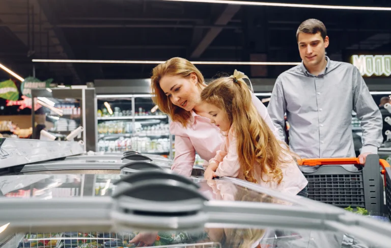 A mother and young daughter lean over a supermarket chest freezer choosing frozen vegetables, while the father follows behind with the trolley.