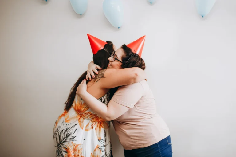 Two women in red party hats share a warm hug, with pale blue balloons on the wall behind them.