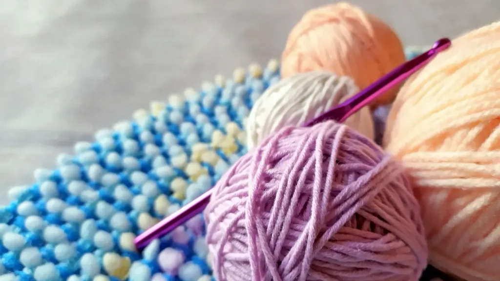 Balls of pastel yarn in lilac, peach and cream with a pink metal crochet hook, resting beside a blue and yellow crocheted piece.