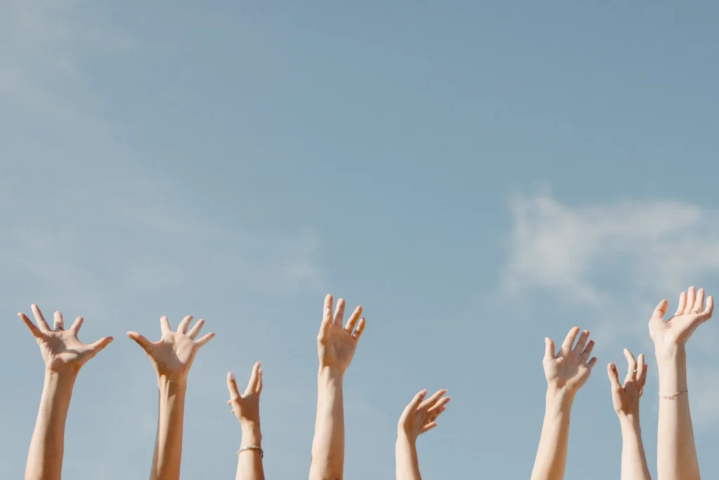 Several raised hands reaching up towards a blue sky with wispy clouds.
