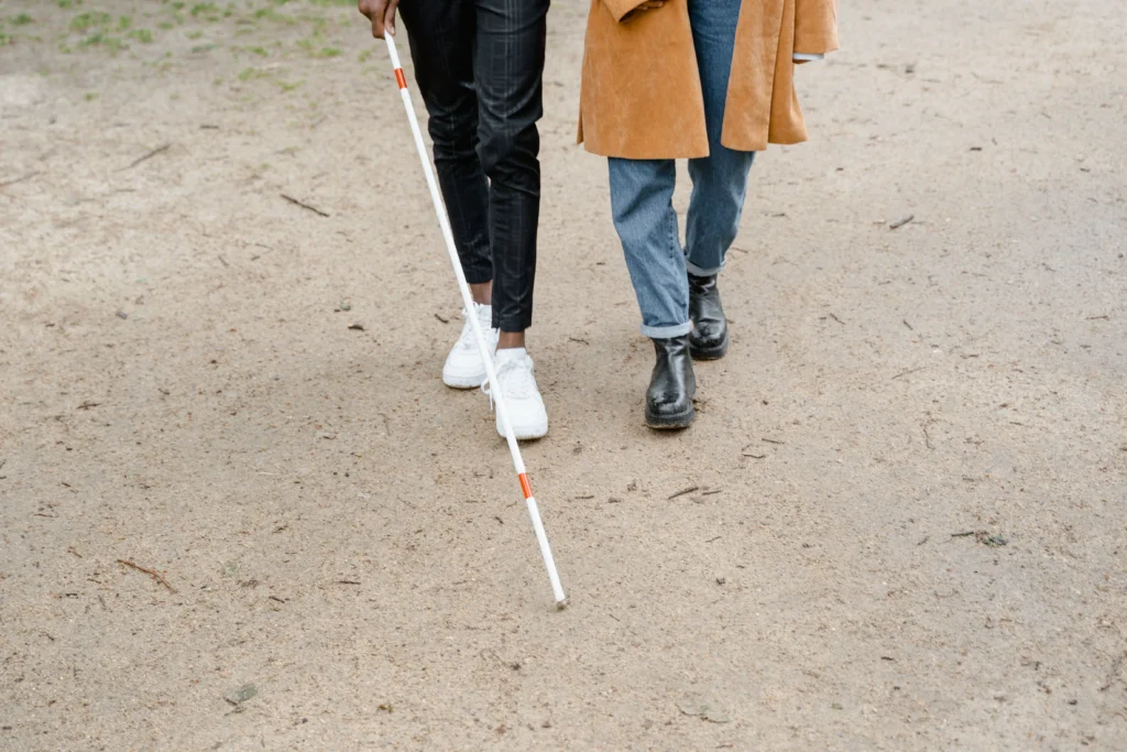 Close-up of two people walking side by side along a gravel path, cropped at the waist — one sweeps a long white cane with red bands across the ground ahead.
