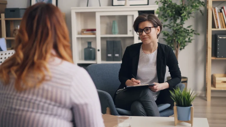 A counsellor in glasses and a dark jacket makes notes on a clipboard while listening to a woman sitting across from her, in a bright, homely office.