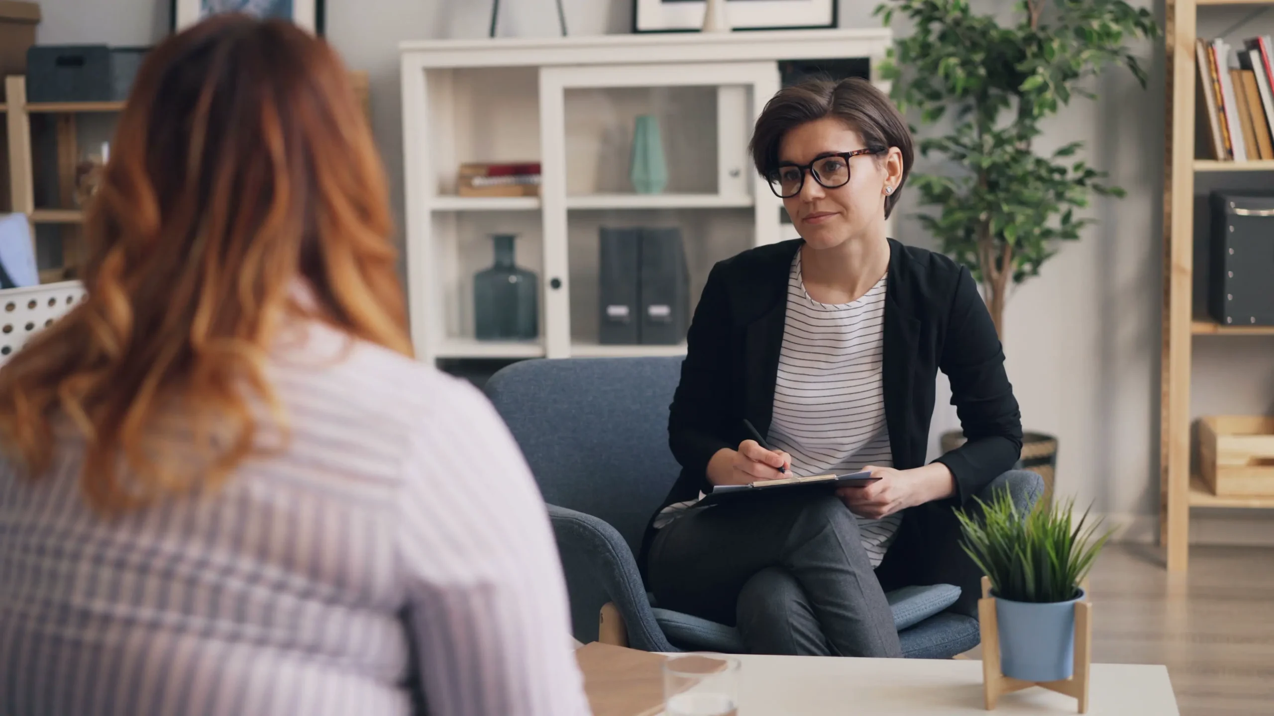 A counsellor in glasses and a dark jacket makes notes on a clipboard while listening to a woman sitting across from her, in a bright, homely office.