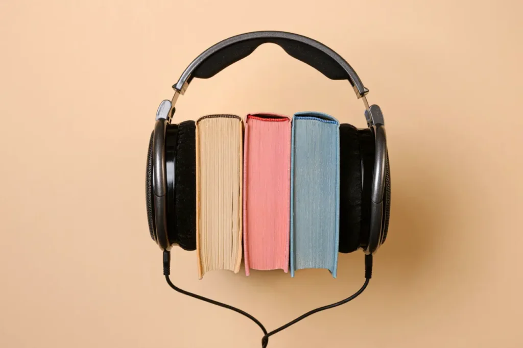 Black over-ear headphones wrapped around three upright books with cream, pink and blue page edges, on a warm beige background — suggesting audiobooks or listening to books.