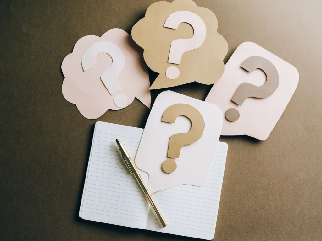 Paper cut-out speech bubbles with question marks in cream and gold, arranged above an open notebook and a gold pen.