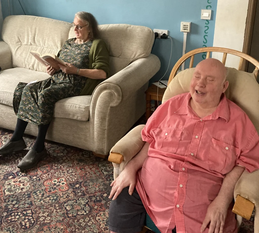 Volunteer and client are seated in a living room with a patterned rug and blue walls. One person sits in an armchair in the foreground wearing a pink shirt, while another sits on a sofa holding and reading from an open book.
