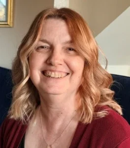Barbara has a warm smile. She has shoulder length wavy auburn hair and is wearing a gold diamond shaped necklace, a black top and maroon cardigan.
