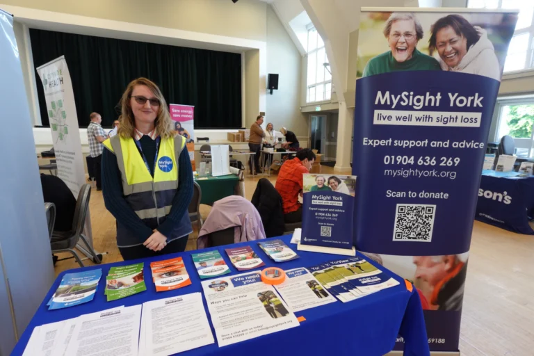 Bex, Mysight York staff member stands in a volunteer vest behind an information stall at a community event, in a large hall.