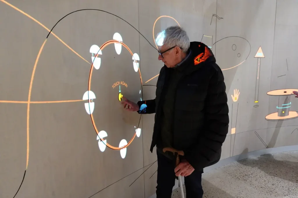 A man holding a white cane reaches out to touch a glowing circular display projected onto a wooden wall.