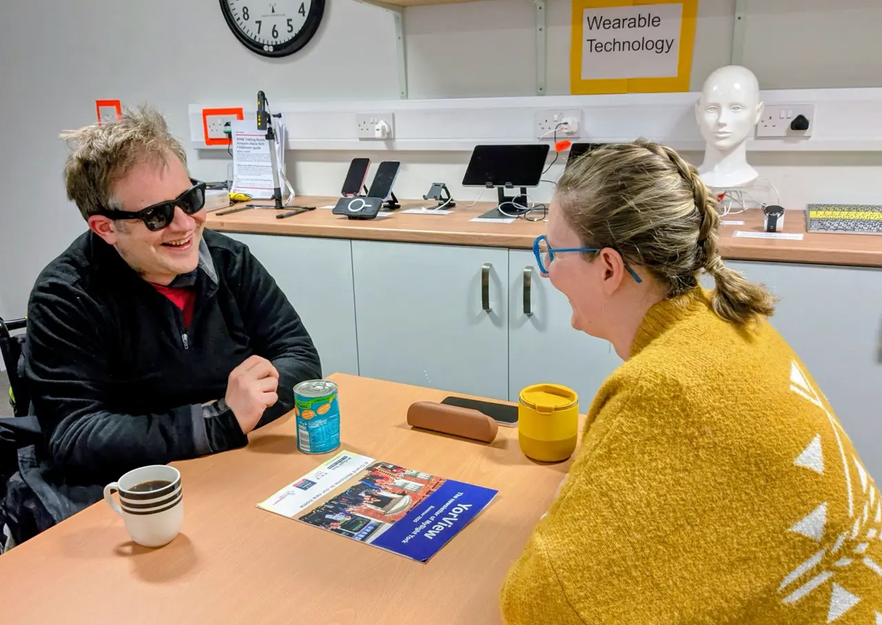 A young man in dark glasses and a woman laugh together across a table, in a room displaying wearable technology.