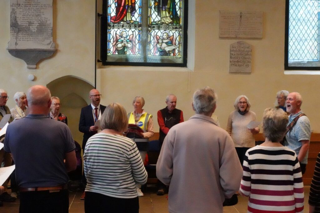 A group of people stand in a circle singing from song sheets in a church, led by a woman at a lectern.