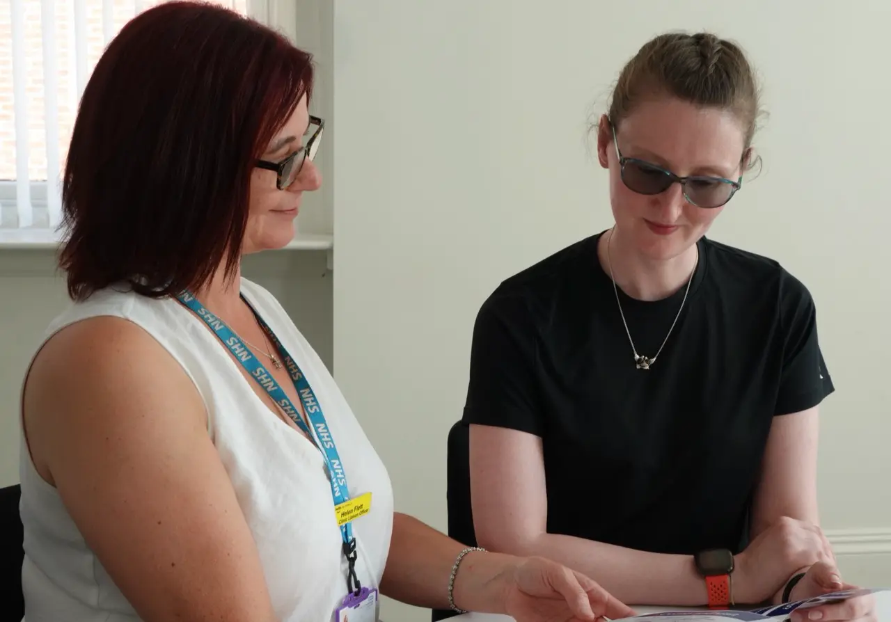 A woman in tinted glasses reads a booklet alongside an eye care liaison officer.