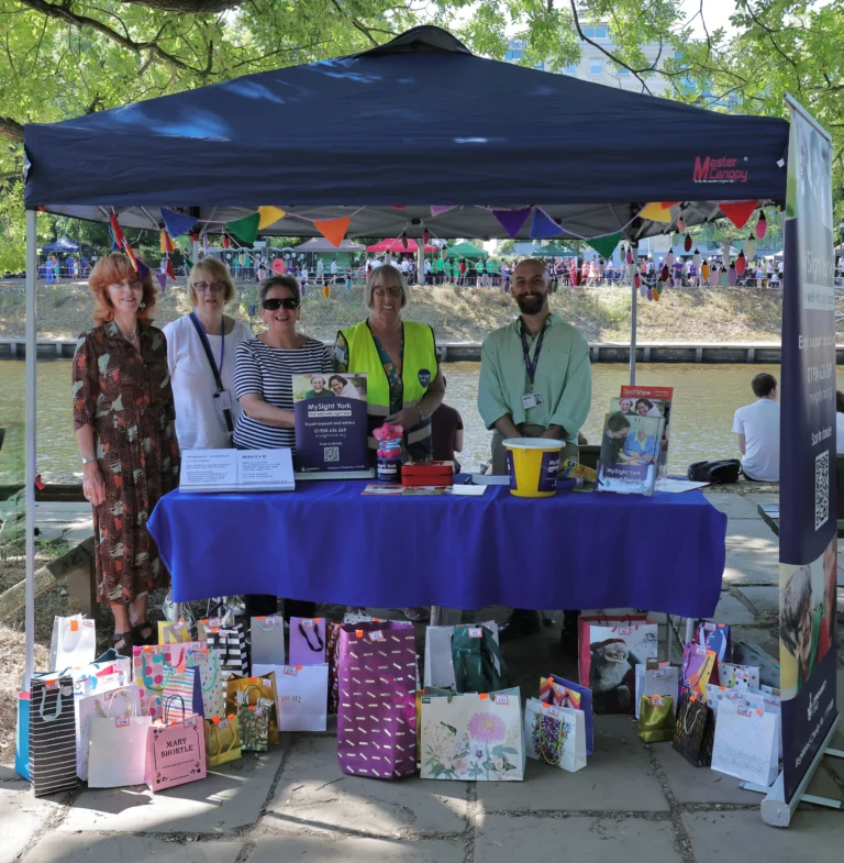 Five volunteers stand behind a MySight York information and fundraising stall beneath a decorated gazebo beside a riverside walkway. The table is covered with a blue cloth and displays promotional materials, donation buckets and raffle information.