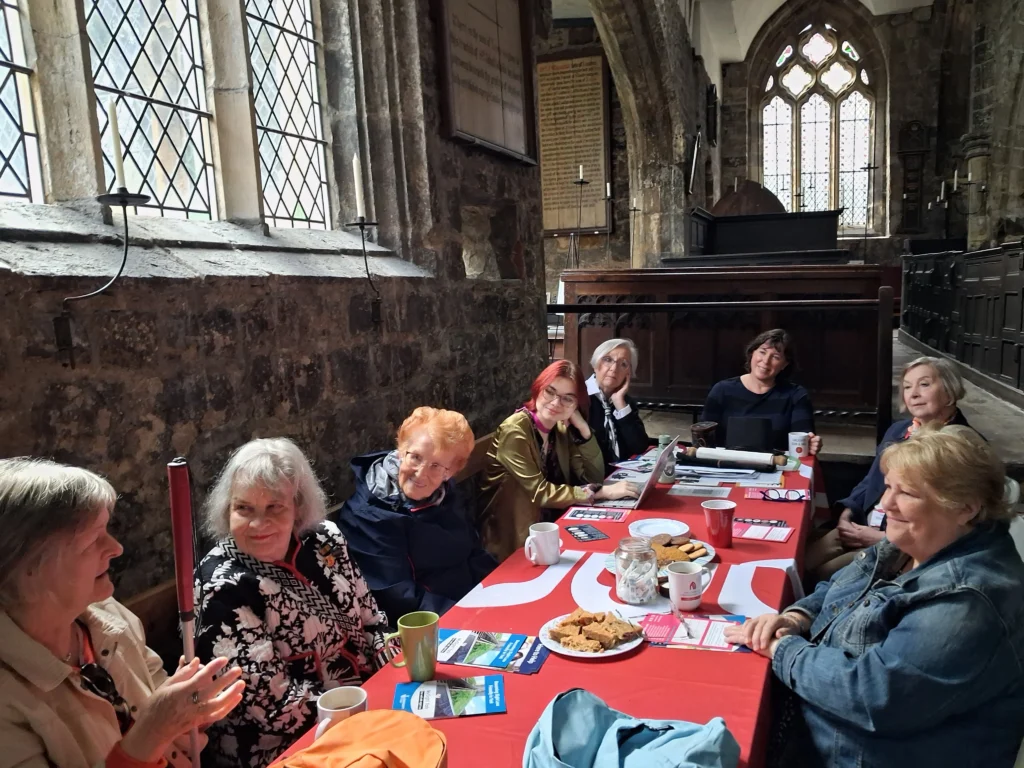 A group of eight people sit around a long table covered with a red tablecloth inside a historic stone church. Drinks, biscuits, leaflets, notebooks and a laptop are spread across the table as participants take part in a consultation meeting.