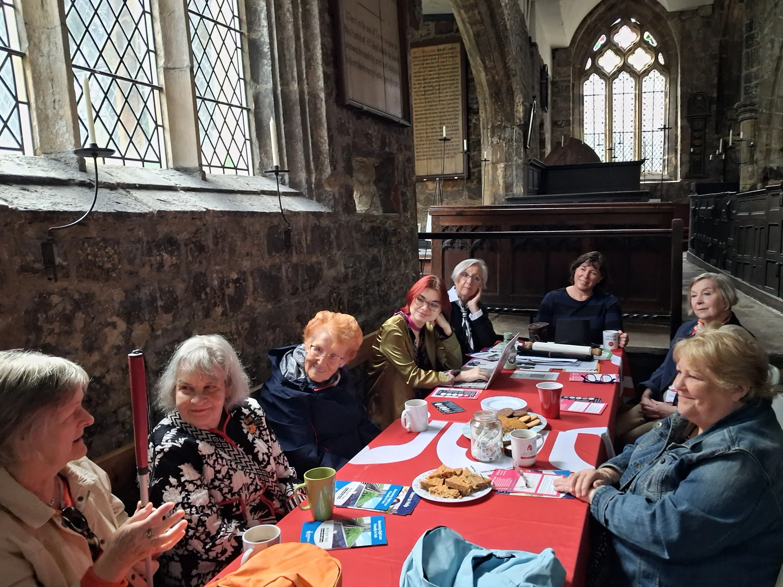 A group of eight people sit around a long table covered with a red tablecloth inside a historic stone church. Drinks, biscuits, leaflets, notebooks and a laptop are spread across the table as participants take part in a consultation meeting.