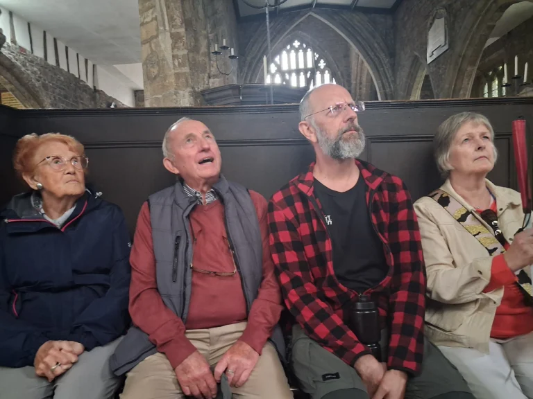 Four people sit side by side on a bench inside a historic stone church, all looking upward toward something out of view, during a consultation.