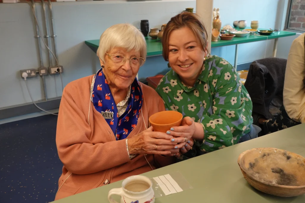A member and staff member sit together at a table during a community event, holding a handmade clay bowl. The table contains pottery items and a mug, while additional ceramic pieces are displayed on shelves in the background.