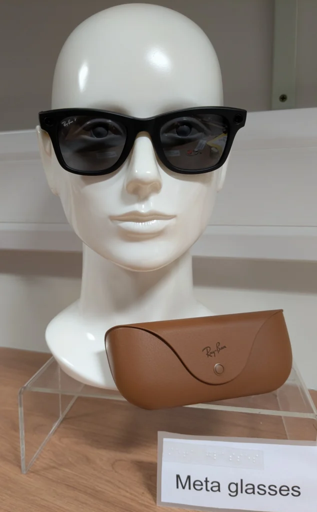 A pair of black smart glasses is displayed on a white mannequin head on a clear acrylic stand. In front of the display is a brown Ray-Ban glasses case, and a label with braille and the text, Meta glasses.