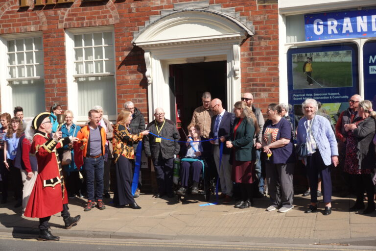 A ribbon-cutting ceremony taking place outside MySight York's building for the Grand Opening. A group of around 20 people stand on the pavement around a blue ribbon stretched across a doorway, while a member in a wheelchair is holding the scissors ready to cut the ribbon.
