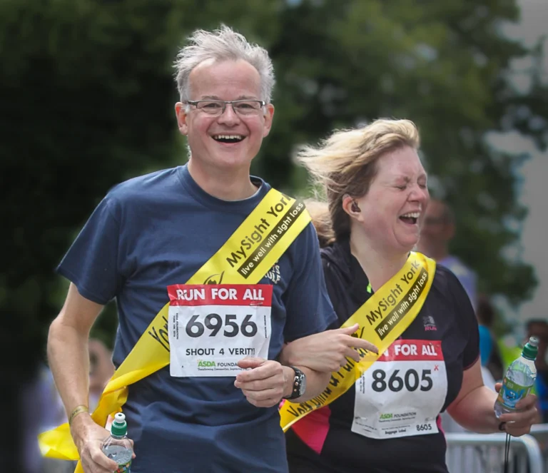 Two participants wearing yellow MySight York sashes take part in a charity running event. Both wear race bibs with the “Run For All” logo and carry water bottles while moving along the course.