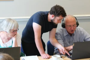Three people sit and stand around a table during a computer learning session. A person in a dark shirt leans over and points at a laptop screen while assisting another person wearing headphones. A third person sits beside them looking at a separate laptop.