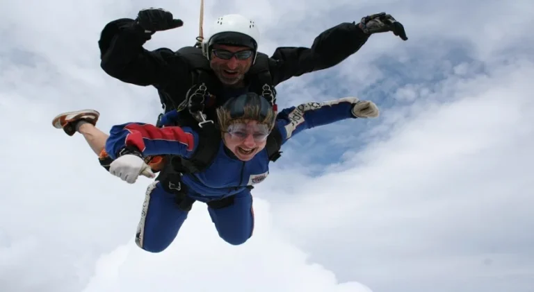 Tandem skydive in progress high above the clouds. Two people are securely harnessed together during freefall, wearing protective helmets, goggles and jumpsuits.