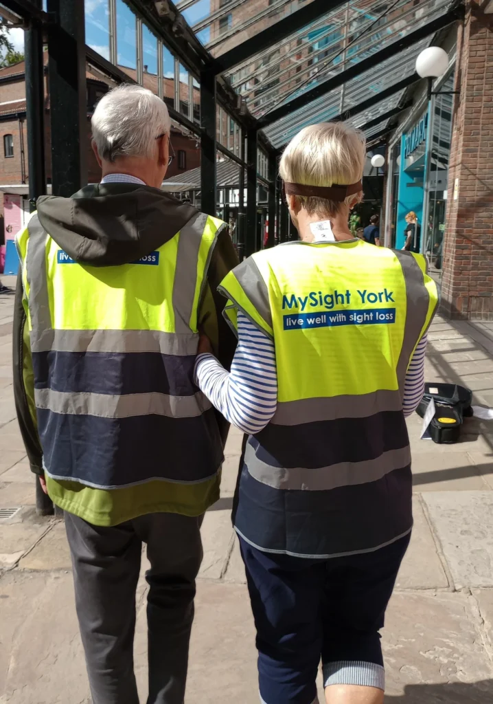 Two volunteers wearing high-visibility MySight York vests walk side by side through a covered pedestrian area. One person holds the other person's arm in a sighted guiding position during a training exercise.