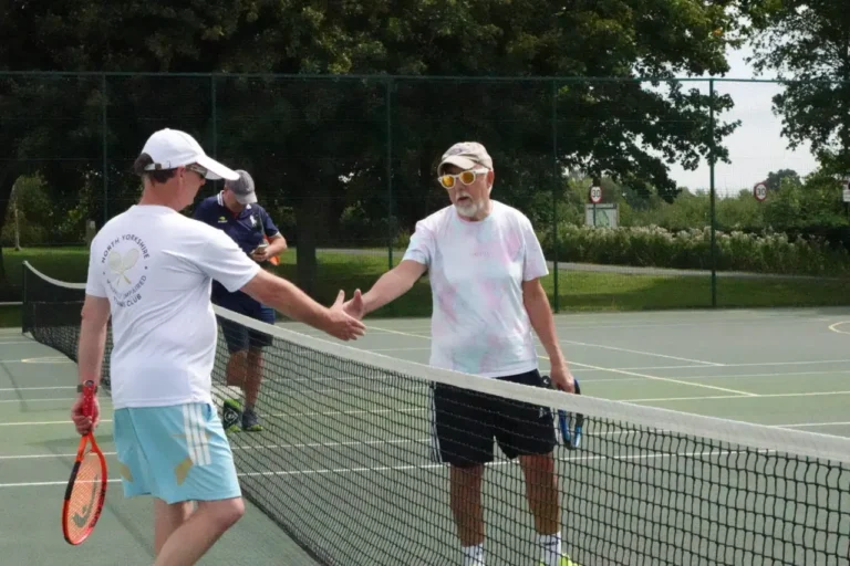 Two men shake hands over a tennis net. One wears a North Yorkshire Visually Impaired Tennis Club shirt, the other dark glasses.