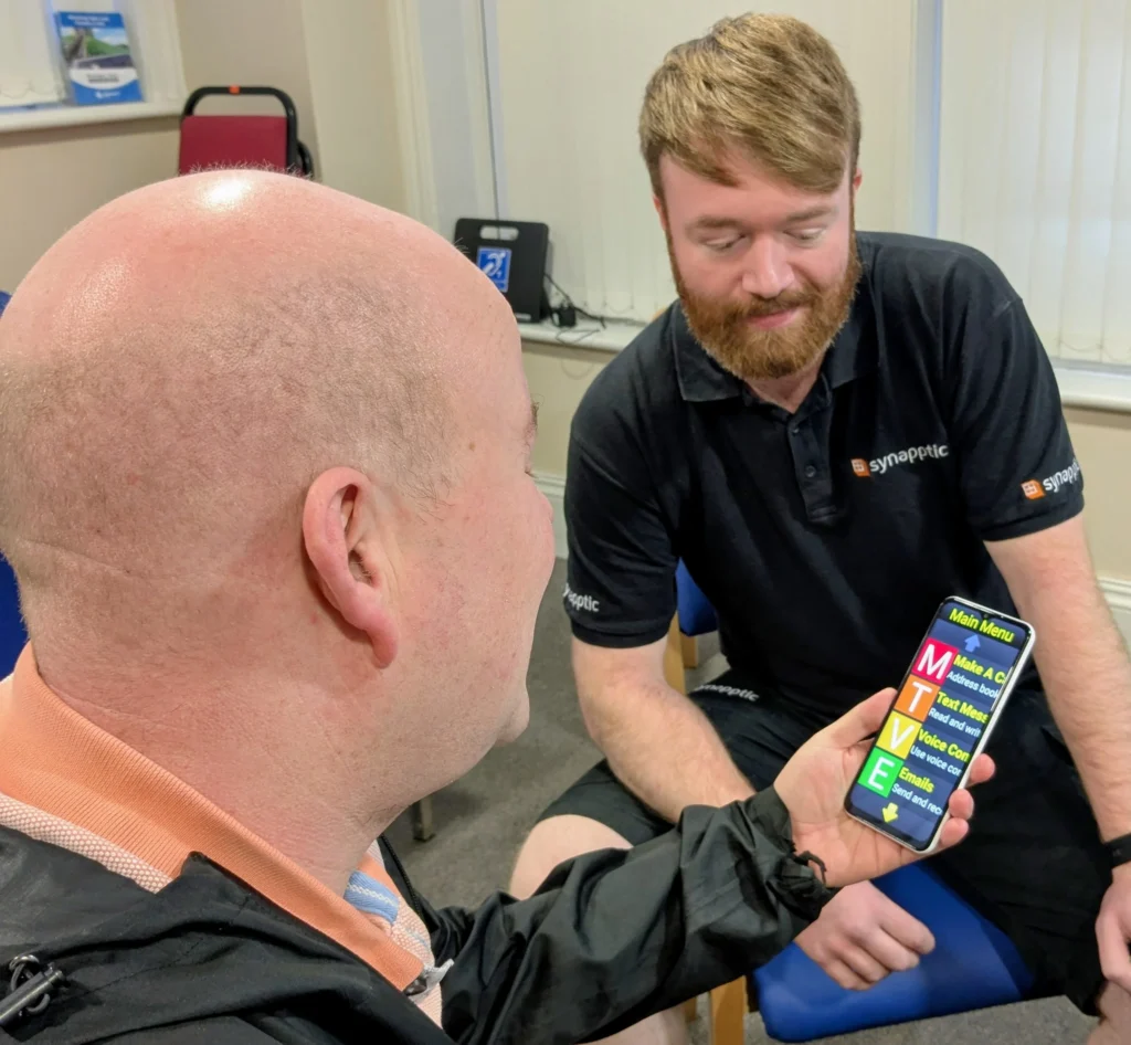 A member seated in a meeting room holds a smartphone displaying a high-contrast, accessible menu with large, colourful buttons for calling, texting, voice controls and email. Another person in a black Synapptic-branded shirt sits nearby, demonstrating the technology.