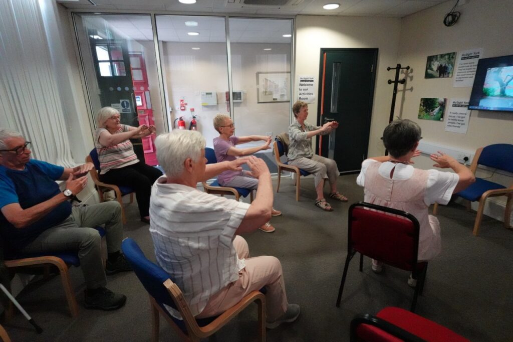 Seven people sit in a circle of chairs in the MySight York activities room, stretching their arms out in front of them.