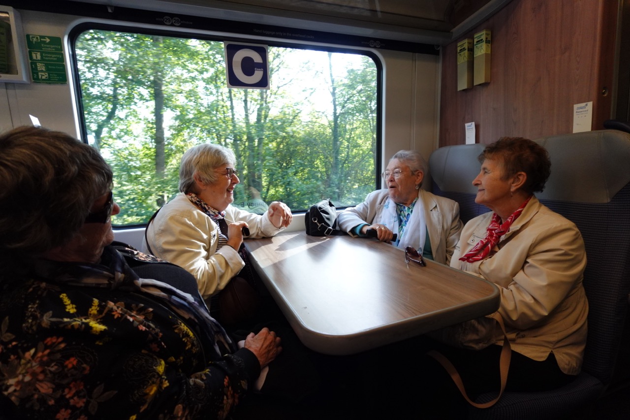 Four women sit around a table on a train, chatting and laughing, with green trees passing the window.