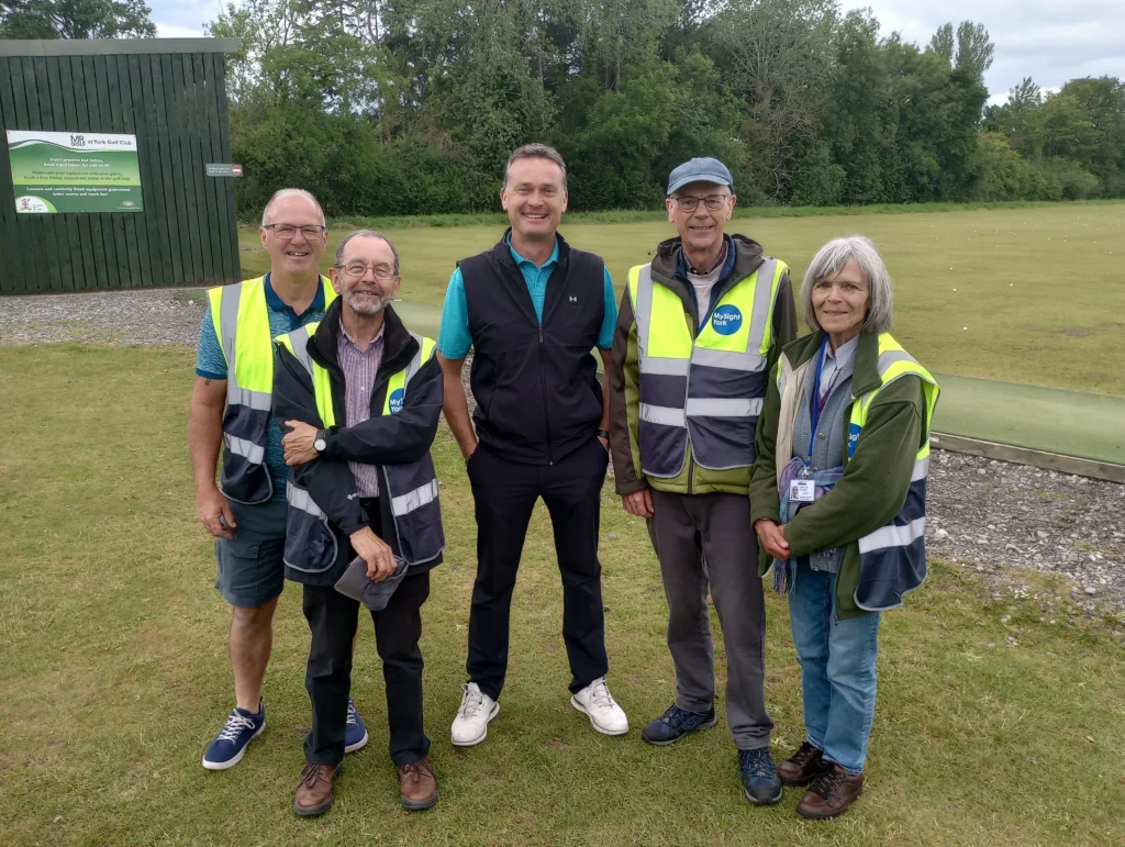 Five people stand together on a golf practice area with grass, trees, and a driving range in the background. Four of the individuals are wearing high-visibility MySight York volunteer vests, while the person in the centre is dressed in golf attire.